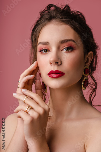 Beauty, fashion portrait of young beautiful woman with pink red color eyes, lips makeup, wearing big trendy earrings, many rings, posing on pink background. 