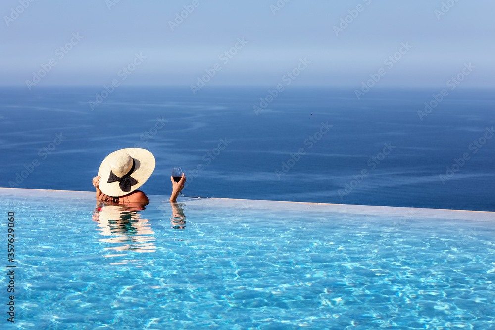 Eine attraktive Frau mit Hut und Weinglas in der Hand genießt die Aussicht vom infinity Pool auf das blaue Meer