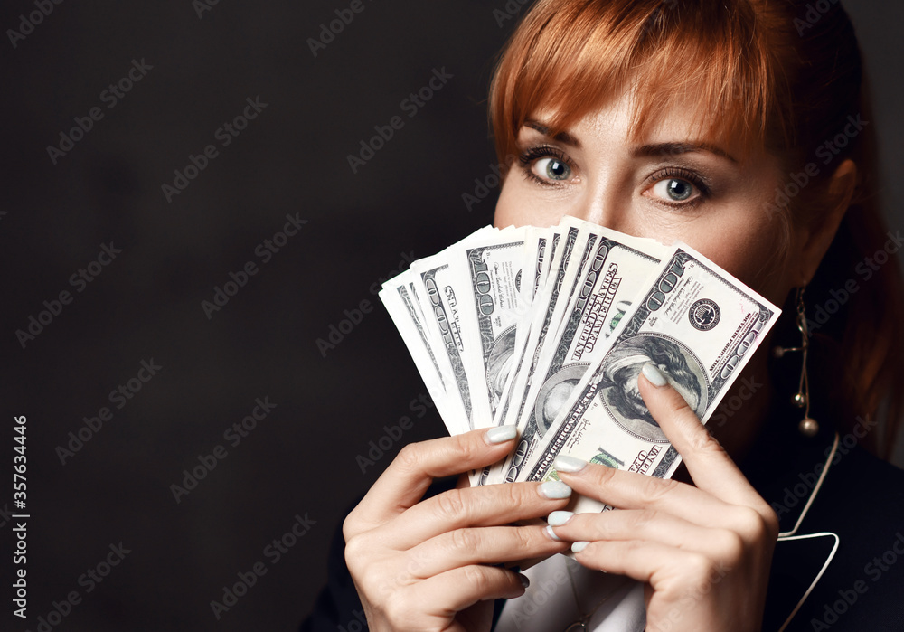 Portrait of young beautiful red-haired business woman in black formal jacket holding heap of dollars in hands Portrait of young beautiful red-haired business woman in black formal jacket holding heap of dollars in hands