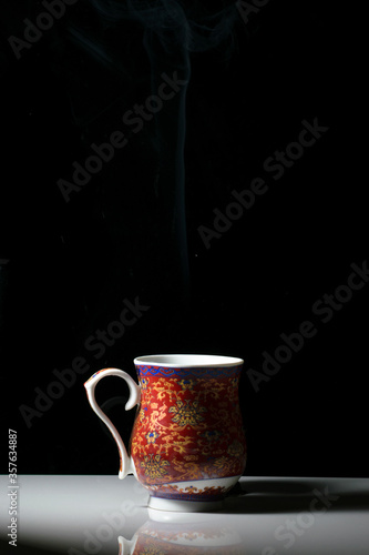 A red tea cup with designs on it placed on a white reflective table and smokes of hot tea coming out of it
