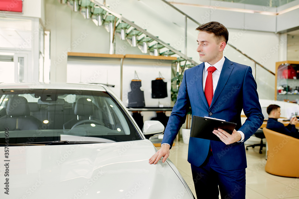 portrait of young affable caucasian consultant of cars dealership standing next to new white car represented for sale portrait of young affable caucasian consultant of cars dealership standing next to new white car represented for sale