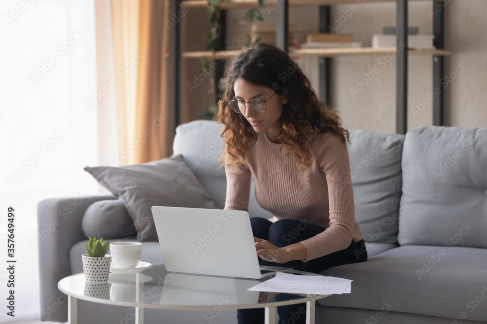 Serious young woman wearing glasses working on laptop, looking at screen, sitting on couch in living room, businesswoman freelancer working on project, writing email, student learning online Serious young woman wearing glasses working on laptop, looking at screen, sitting on couch in living room, businesswoman freelancer working on project, writing email, student learning online