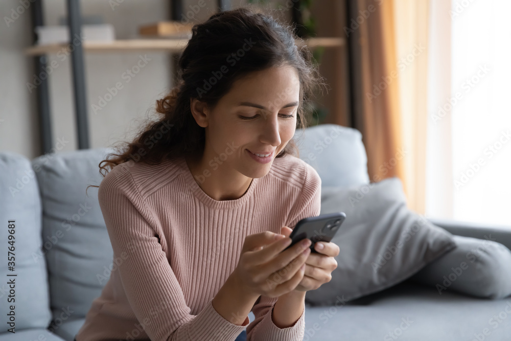 Smiling young woman using smartphone close up, sitting on cozy couch in living room, beautiful girl holding phone, looking at screen, chatting in social network with friends, browsing apps Smiling young woman using smartphone close up, sitting on cozy couch in living room, beautiful girl holding phone, looking at screen, chatting in social network with friends, browsing apps