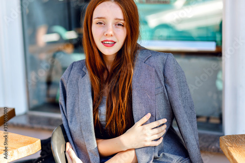 Outdoor fashion portrait of stylish ginger woman, posing at terrace cafe, at sunny day, wearing boyfriend jacket, bright fresh colors.


 photo