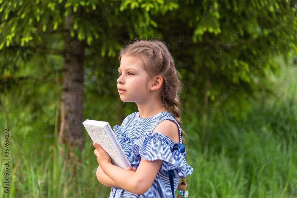 A beautiful little girl holds a book in a green forest and looks into the distance. A beautiful little girl holds a book in a green forest and looks into the distance.