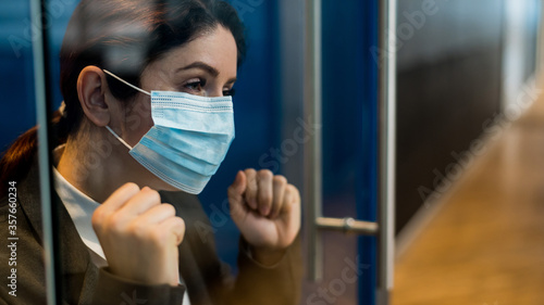 A girl in a medical mask is isolated due to biological hazard. The business woman in the suit is quarantined and leaned against the glass door with sadness. The epidemic of the virus.