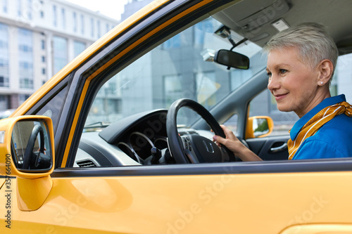 Beautiful mature female with pixie hair driving car, enjoying empty streets early in the morning. Attractive middle aged woman sitting in driver's seat, parking automobile, looking at side view mirror