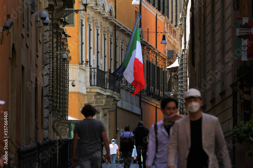 Street in the historic center of Rome. Recovery phase after the lockdown. Italian flag on the background, people walking with face mask.
