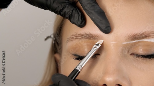 Master applies brow paste with a brush to eyebrows. Beautiful attractive female face of a blonde well-groomed woman or lady. Styling and lamination of eyebrows. Stylist's hands in black gloves