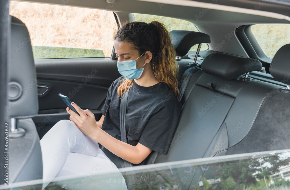 Naklejka premium New normal. Teenager sitting in the back of a car with a mask. Young woman in a taxi protected by a mask. Safety and pandemic concept. Coronavirus devices. Social distance. Young girl using smartphone