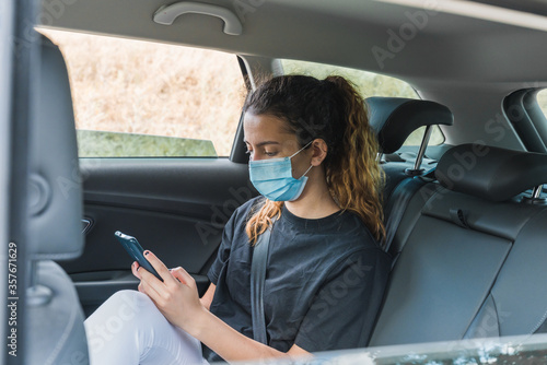 New normal. Teenager sitting in the back of a car with a mask. Young woman in a taxi protected by a mask. Safety and pandemic concept. Coronavirus devices. Social distance. Young girl using smartphone