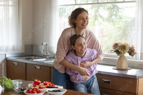 Overjoyed young Caucasian mother and happy teenage daughter have fun, joke and laugh cooking together, playful mom and smiling teen girl child feel crazy entertain preparing food in home kitchen
