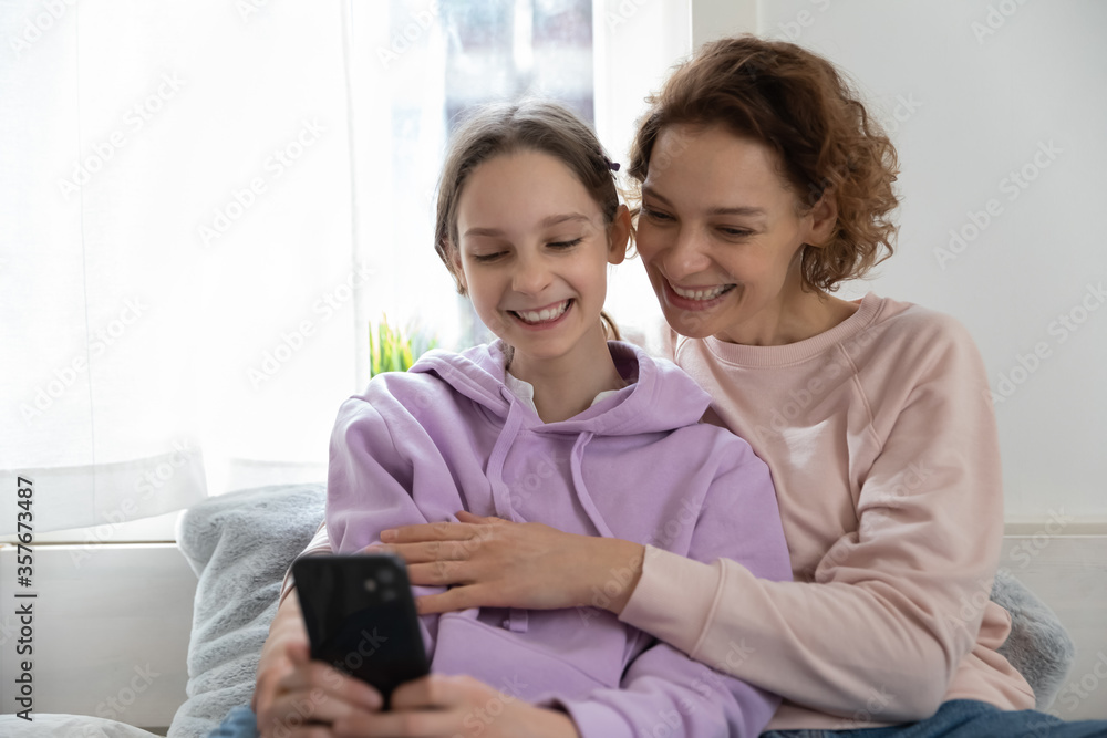 Smiling young mother and teenage daughter make self-portrait picture on smartphone together, happy mom and teen girl have fun using playing on cellphone gadget at home, technology concept Smiling young mother and teenage daughter make self-portrait picture on smartphone together, happy mom and teen girl have fun using playing on cellphone gadget at home, technology concept