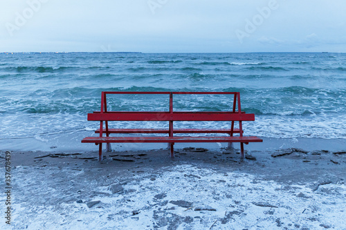 Red bench covered by ice is hit by winter sea. Icy waves of Baltic sea on empty beach.