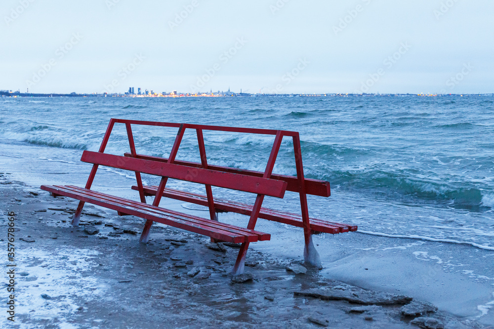 Red bench covered by ice is hit by winter sea. Icy waves of Baltic sea on empty beach.