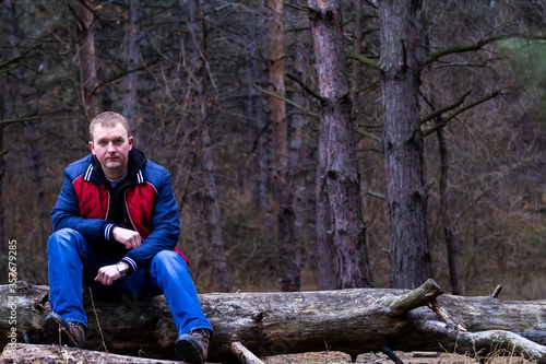 young attractive man dressed in a red jacket with blue sleeves sits on a log