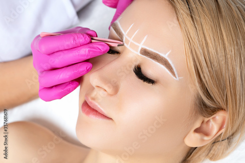Woman Having Permanent Make-up Tattoo on her Eyebrows. Eyelash maker plucks eyebrows with tweezers. Professional makeup and cosmetology skin care.