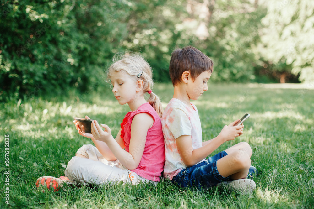 Boy and girl play games on smartphones outdoors. Kids digital gadget screen addiction. Children friends playing online sitting on grass in park. Contemporary problem of loneliness together. Boy and girl play games on smartphones outdoors. Kids digital gadget screen addiction. Children friends playing online sitting on grass in park. Contemporary problem of loneliness together.