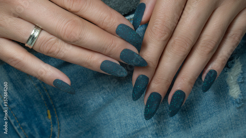 Blue manicure with a glitter. Dark blue glitter nails on jeans textile background.