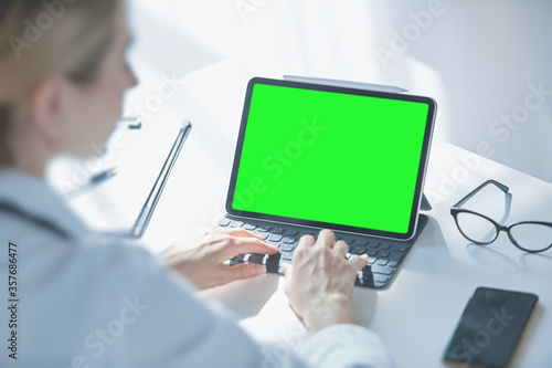 Medical worker a woman in a white coat uses a tablet in the office at the Desk, chromakey on the tablet screen, a view over her shoulder
