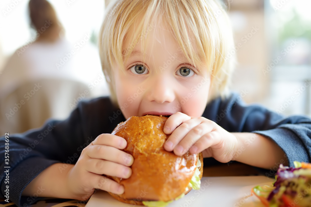 Cute blonde boy eating large hamburger at fastfood restaurant. Unhealthy meal for kids. Junk food. Cute blonde boy eating large hamburger at fastfood restaurant. Unhealthy meal for kids. Junk food.