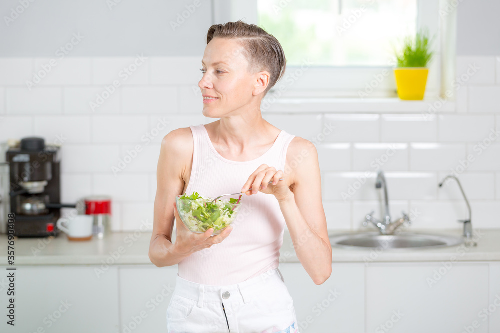 Young woman eating healthy food sitting in the beautiful white kitchen interior with green flowers on the background Young woman eating healthy food sitting in the beautiful white kitchen interior with green flowers on the background