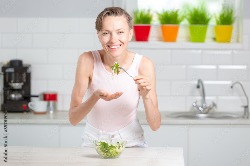 Young woman eating healthy food sitting in the beautiful white kitchen interior with green flowers on the background Young woman eating healthy food sitting in the beautiful white kitchen interior with green flowers on the background