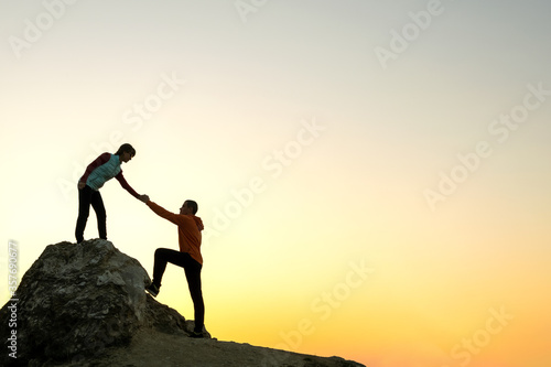 Man and woman hikers helping each other to climb a big stone at sunset in mountains. Couple climbing on a high rock in evening nature. Tourism, traveling and healthy lifestyle concept.
