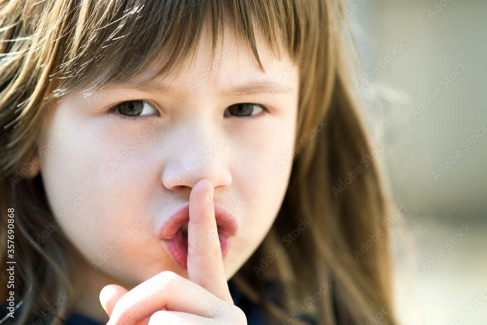 Portrait of pretty child girl with gray eyes and long hair holding point finger on her lips with hush sigh. Cute female kid on warm summer day outside. Portrait of pretty child girl with gray eyes and long hair holding point finger on her lips with hush sigh. Cute female kid on warm summer day outside.