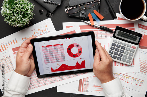 Top view of businessman's hands working with tablet pc and financial reports. Modern black office desk with notebook, pencil and a lot of things. Flat lay table layout.