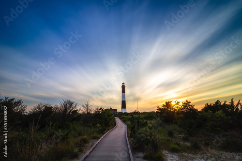 Wooden boardwalk leading to a tall stone lighthouse  as sunset clouds streak across the sky overhead. Fire Island New York
