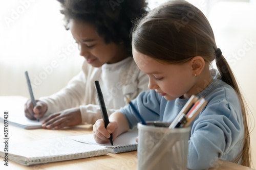 African Caucasian schoolgirl mates sit at desk writing task learning subject in classroom. Multiracial girls use felt-tip pen noting on workbook do schoolwork at home. Homeschooling, education concept photo