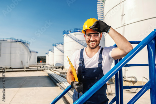 Young hardworking Caucasian unshaven refinery worker in working clothes with helmet on head and protective glasses holding folder with documents under armpit and looking at camera.
