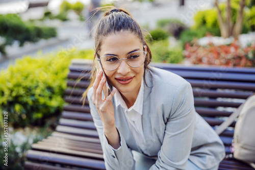 Young attractive brunette with eyeglasses dressed smart casual sitting on the bench in park and having phone conversation.