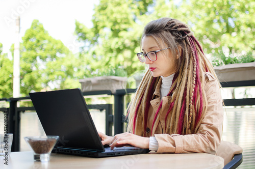 Skilled charming female freelancer with long dreadlocks hair is using black laptop computer during a distance work outside sitting at the table in street coffee shop outside in spring or autumn