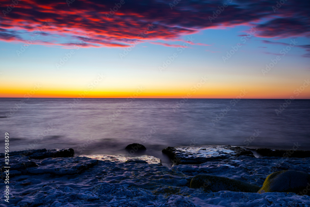 Obraz premium Vibrant colored summer sunset reflecting in ocean with endless horizon over deep blue ocean, silhouette of boulders that is laying in the foreground in shallow water at island of Gotland, Sweden