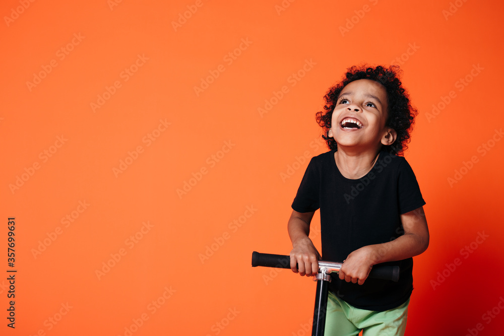 Portrait of an African American boy who throws his head back and riding a scooter in a studio on an orange background. Portrait of an African American boy who throws his head back and riding a scooter in a studio on an orange background.