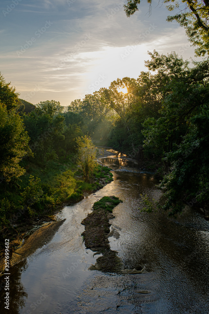 Sun rising shining through green leaves tree forest over a stream river landscape in Girona, Catalonia Sun rising shining through green leaves tree forest over a stream river landscape in Girona, Catalonia
