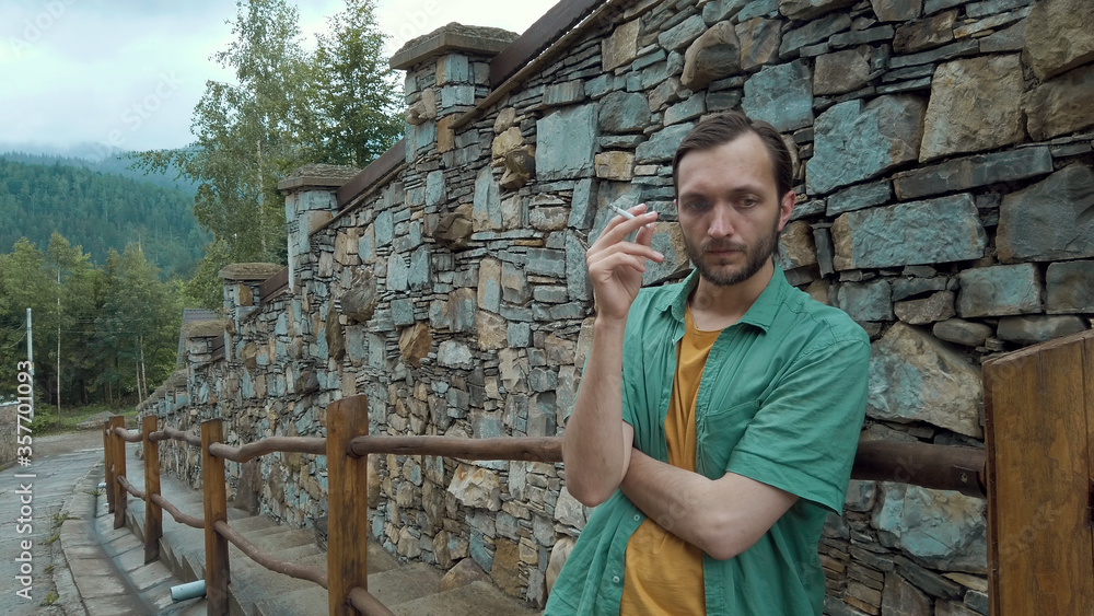 Depressed man smoking cigarette, feeling disappointed, standing along the road, stone wall on background. Male with bad habit.