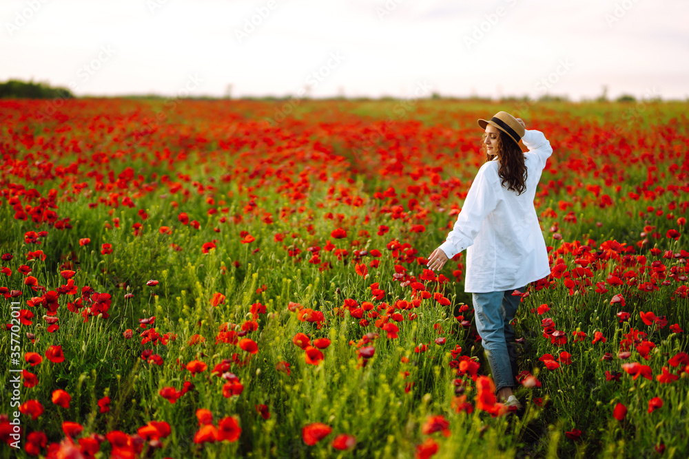 Fototapeta premium Young girl in hat posing in the poppy field. Portrait of curly girl walking in poppy field at sunset. Summertime.
