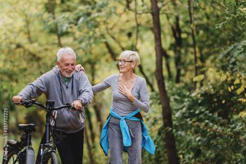 Cheerful active senior couple with bicycles walking through park together. Perfect activities for elderly people. © lordn