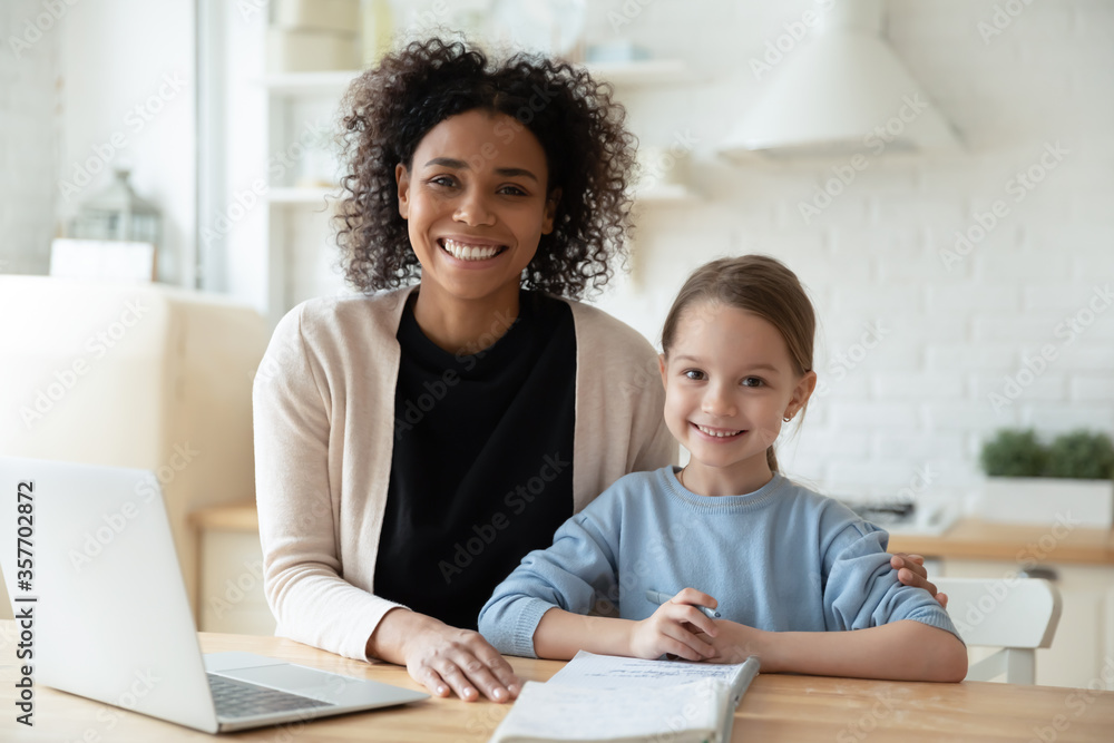 Happy African female tutor and little learner schoolgirl portrait. Multi-racial family sit in kitchen laptop and workbooks lie on table. Successful homeschooling, clever kid girl study at home concept Happy African female tutor and little learner schoolgirl portrait. Multi-racial family sit in kitchen laptop and workbooks lie on table. Successful homeschooling, clever kid girl study at home concept