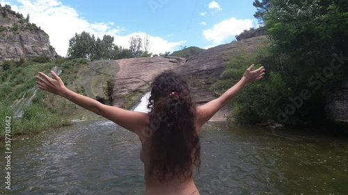 Nudist girl from the back raises her arms in a river and a waterfall in a natural landscape photo