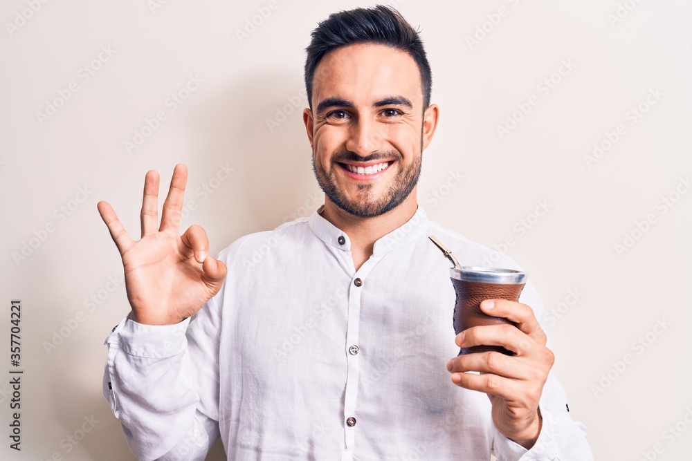 Young handsome man with beard drinking mate infusion beverage over white background doing ok sign with fingers, smiling friendly gesturing excellent symbol Young handsome man with beard drinking mate infusion beverage over white background doing ok sign with fingers, smiling friendly gesturing excellent symbol