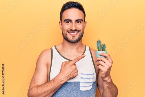 Young handsome man with beard wearing sleeveless t-shirt holding cactus plant pot smiling happy pointing with hand and finger