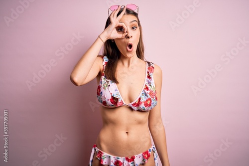 Young beautiful woman on vacation wearing bikini and sunglasses over pink background doing ok gesture shocked with surprised face, eye looking through fingers. Unbelieving expression. © Krakenimages.com