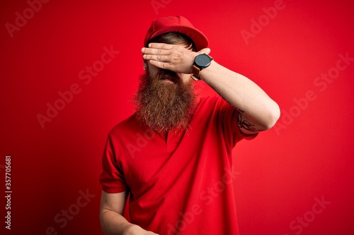 Young handsome delivery man wearing glasses and red cap over isolated background covering eyes with hand, looking serious and sad. Sightless, hiding and rejection concept © Krakenimages.com