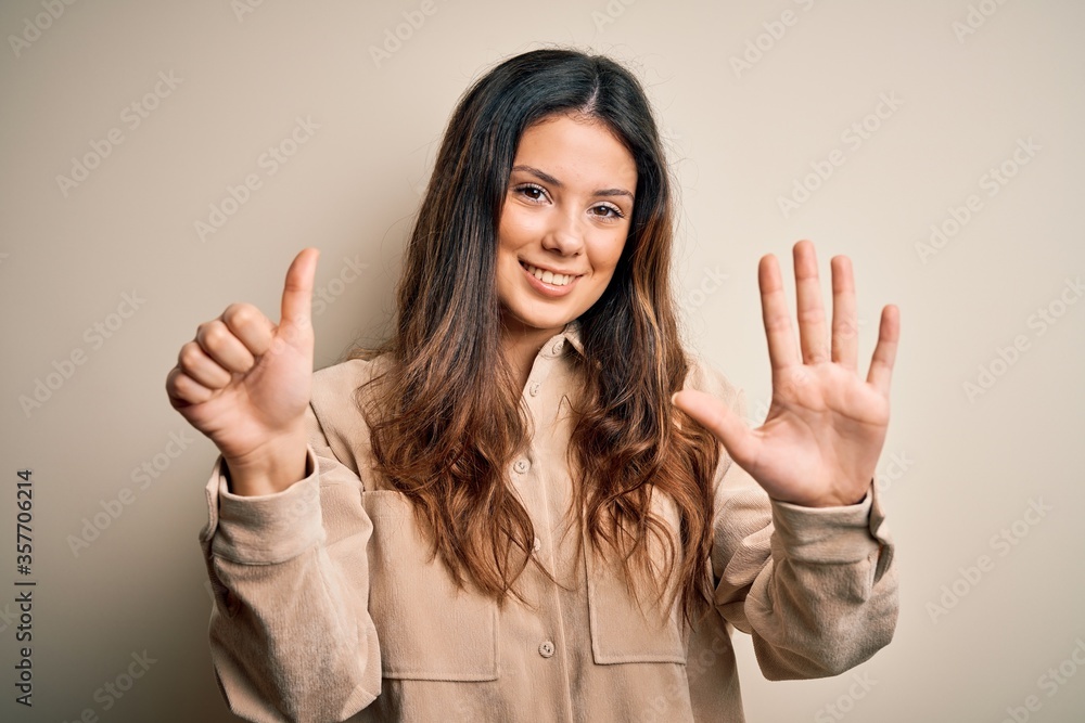 Young beautiful brunette woman wearing casual shirt standing over white background showing and pointing up with fingers number six while smiling confident and happy. Young beautiful brunette woman wearing casual shirt standing over white background showing and pointing up with fingers number six while smiling confident and happy.