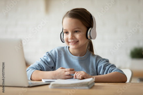 Wearing wireless headphones makes videocall little beautiful schoolgirl using web cam and notebook learning school subject distantly due quarantine or home schooling. Remote self-education concept photo