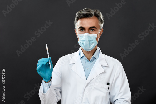 Mature doctor or scientist in uniform and medical mask holding syringe with vaccine from flu, coronavirus, covid-19 while standing against black background